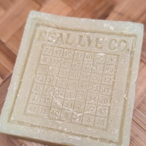 May include: A square, light green bar of soap with the text "REAL LYE CO." and a grid of numbers. The soap has a textured surface and a rectangular border. The numbers are arranged in a grid pattern.