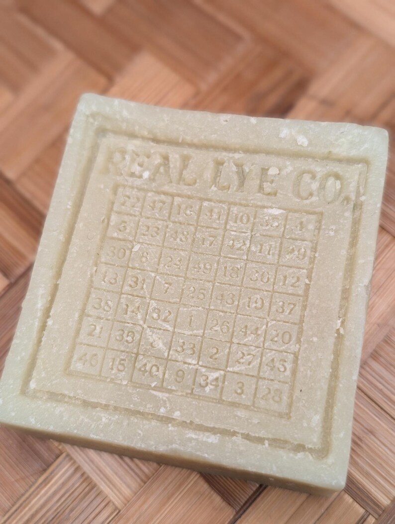 May include: A square, light green bar of soap with the text "REAL LYE CO." and a grid of numbers. The soap has a textured surface and a rectangular border. The numbers are arranged in a grid pattern.