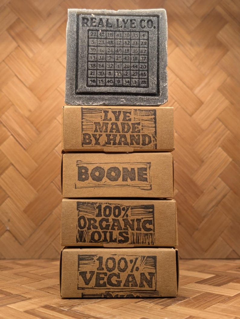 May include: A stack of four brown cardboard boxes with black text and a gray bar of soap. The top box reads "LYE MADE BY HAND". The soap is labeled "REAL LYE CO." and "100% VEGAN".