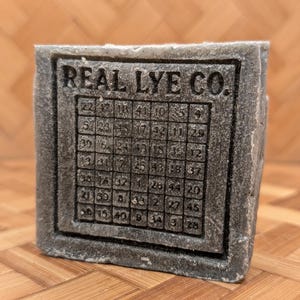 May include: A gray soap bar with the text "REAL LYE CO." and a grid of numbers. The soap has a textured appearance and a rectangular shape. The numbers are arranged in a square pattern.