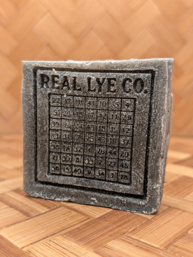 May include: A gray soap bar with the text "REAL LYE CO." and a grid of numbers. The soap has a textured appearance and a rectangular shape. The numbers are arranged in a square pattern.