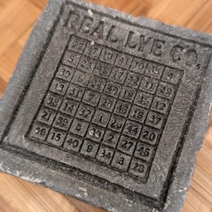 May include: A gray, rectangular soap mold with the text "REAL LYE CO." and a grid of numbers. The numbers are arranged in a square pattern, likely for soap making. The mold has a textured surface.