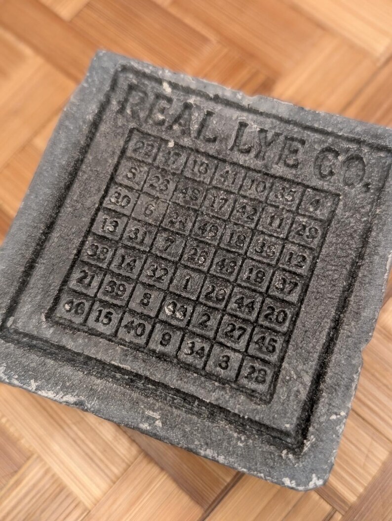 May include: A gray, rectangular soap mold with the text "REAL LYE CO." and a grid of numbers. The numbers are arranged in a square pattern, likely for soap making. The mold has a textured surface.