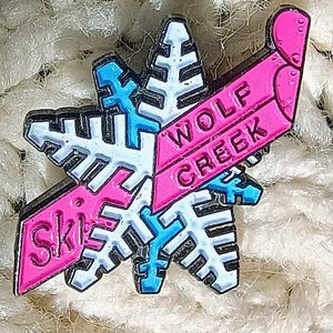 May include: A snowflake-shaped pin with the words "Ski" and "Wolf Creek" in pink and white. The pin has a blue and white snowflake design. The pin is set against a textured, off-white background.