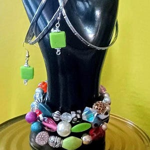 May include: A pair of silver-toned hoop earrings featuring lime green square beads and a multi-strand bracelet composed of assorted colorful beads, including pearls, faceted crystals, and textured metallic spheres, displayed on a black hand form.