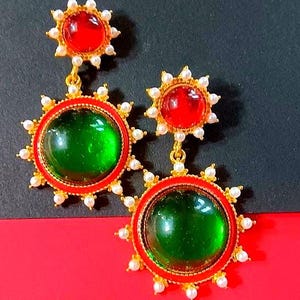 May include: A pair of gold-toned earrings with red and green gemstone accents. The earrings feature a sunburst design with small white pearls around the gemstones. The background is red and black.
