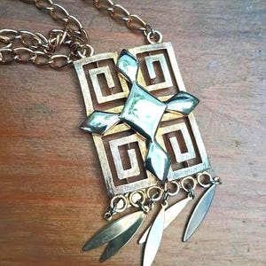 May include: A gold-tone necklace featuring a rectangular pendant with a cut-out Greek key pattern. A central diamond-shaped element sits atop the frame, and four small, smooth, leaf-shaped charms dangle from the bottom edge of the pendant.