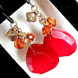 May include: A pair of dangle earrings featuring large, faceted red teardrop-shaped stones. The earrings have gold-colored metal accents, including small beads and geometric charms. The ear wires are dark-colored.