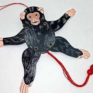 May include: A wooden monkey toy with articulated limbs, painted black with white accents. The monkey has a light brown face and pink hands and feet. A red string is attached to the monkey's body and tail, with red beads at the end.