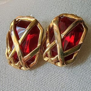 May include: A pair of vintage clip-on earrings. Each earring features a rectangular, faceted, red gemstone encased in a gold-tone metal cage design. The earrings are set against a neutral background.