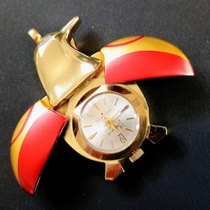 May include: A gold-toned ladybug-shaped watch with red and yellow wings. The watch face is silver with gold hands and markers. The watch is on a dark surface.