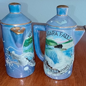 May include: Two blue ceramic teapots with gold trim and handles. One teapot features a dragon design, the other depicts Niagara Falls with the text "Niagara Falls Prospect Point". These decorative teapots are likely souvenirs.