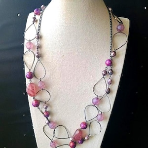 May include: A layered necklace with a mix of pink and purple beads and teardrop-shaped metal accents. The necklace is displayed on a white mannequin, showcasing its design and color variations. The beads vary in size and shape.