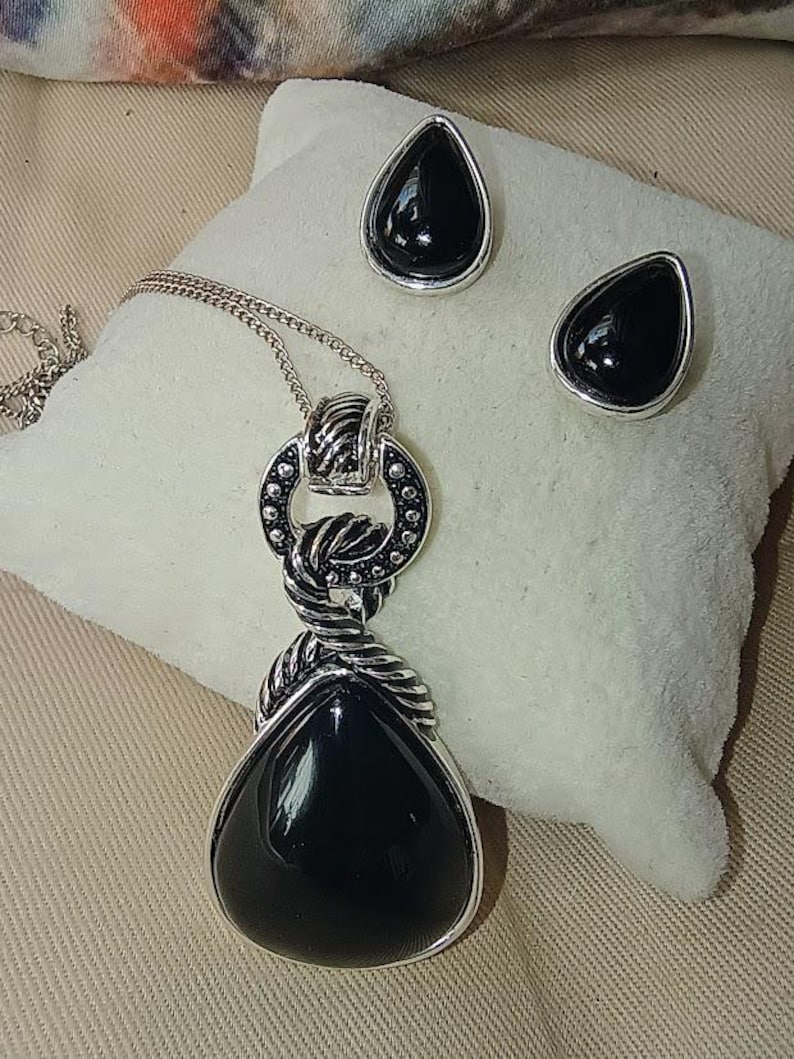 以下が含まれることがあります： シルバーカラーのジュエリーセット。涙型のペンダントネックレスと、お揃いのスタッドピアスが特徴です。ペンダントとピアスは、黒く光沢のある石がシルバーのフレームにセットされています。ネックレスチェーンは繊細です。