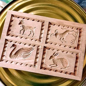May include: A light brown wooden mold with four carved animal designs: a goat, a ram, a swan, and a rabbit. The mold is set on a gold-colored tray. The carvings are detailed and the wood has a natural finish.