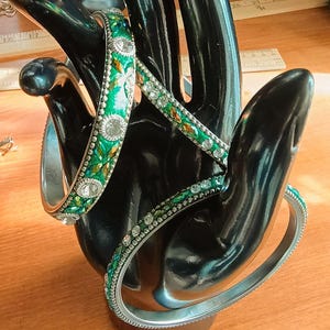 May include: Several green and silver bangles with jewel and rhinestone embellishments are displayed on a black hand-shaped jewelry holder. The bangles have a variety of designs, including floral and geometric patterns.