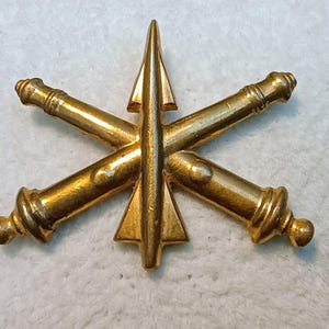 May include: A gold-colored military insignia featuring crossed cannons and a central missile or arrow. The emblem has a detailed, textured surface and a polished finish, suggesting a vintage or commemorative item.