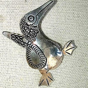 May include: A silver-toned bird brooch with intricate detailing. The brooch features a bird with a long beak, detailed wings, and webbed feet. The bird's head and wings have textured patterns. The brooch is likely made of metal and is a decorative accessory.