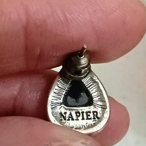 以下が含まれることがあります： 黒い中心部を持つ、銀色の涙の形をしたイヤリング。 イヤリングにはピアス用のポストがあり、下部には「NAPIER」の文字が刻まれています。 イヤリングは指で持たれています。