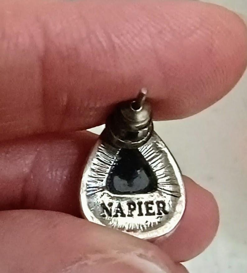 以下が含まれることがあります： 黒い中心部を持つ、銀色の涙の形をしたイヤリング。 イヤリングにはピアス用のポストがあり、下部には「NAPIER」の文字が刻まれています。 イヤリングは指で持たれています。