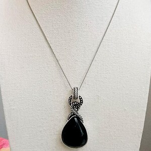 以下が含まれることがあります： 黒いしずく型のペンダントが付いたシルバーカラーのネックレス。ペンダントはロープのようなデザインの装飾的なベイルから吊り下げられています。ネックレスは白いマネキンに展示されています。