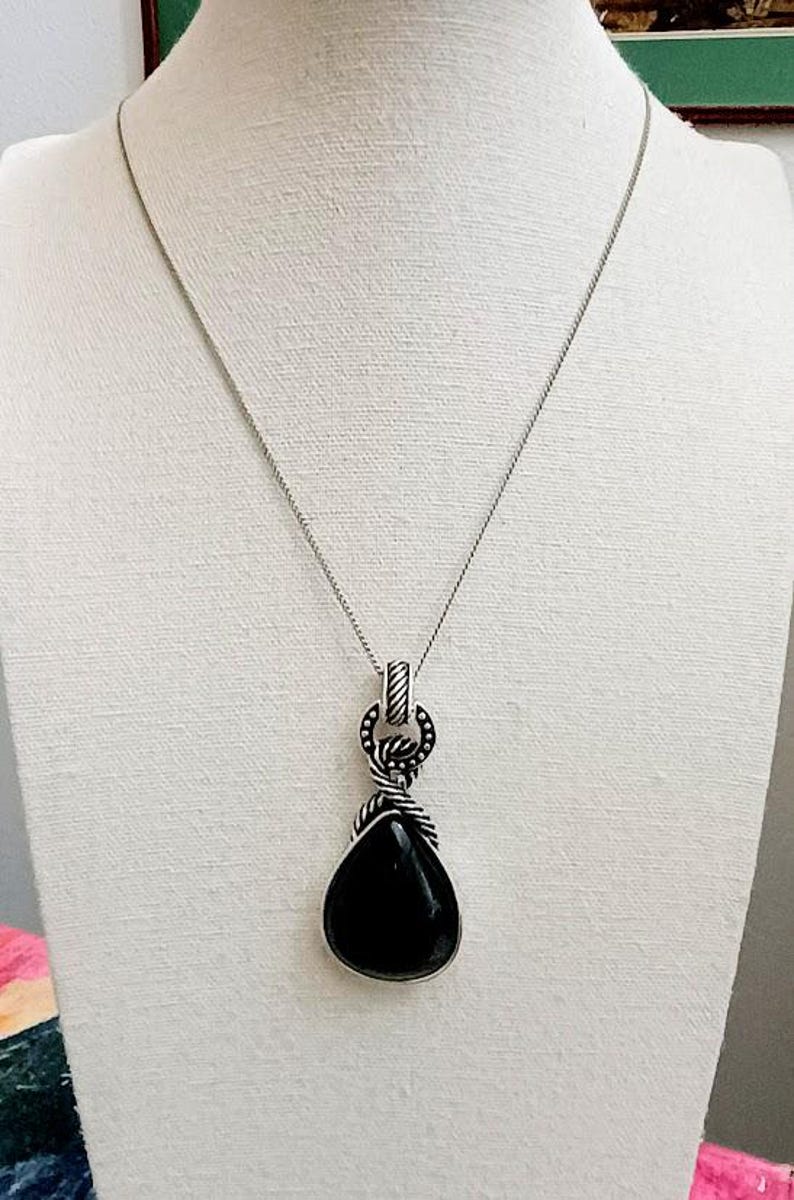 以下が含まれることがあります： 黒いしずく型のペンダントが付いたシルバーカラーのネックレス。ペンダントはロープのようなデザインの装飾的なベイルから吊り下げられています。ネックレスは白いマネキンに展示されています。