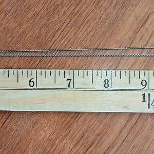 以下が含まれることがあります： ハート型のペンダントが付いたシルバーカラーのネックレス。ペンダントの幅は約2.5cm、高さは約2.5cmです。ネックレスは木製の定規の上に置かれており、チェーンは左に伸びています。定規はインチで表示されています。