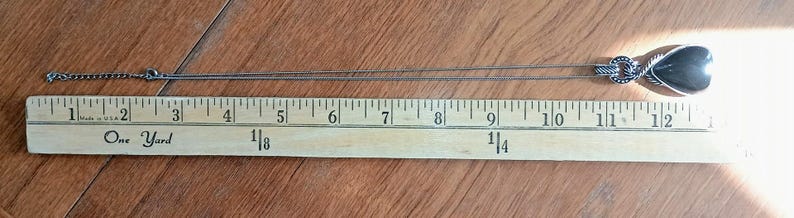 以下が含まれることがあります： ハート型のペンダントが付いたシルバーカラーのネックレス。ペンダントの幅は約2.5cm、高さは約2.5cmです。ネックレスは木製の定規の上に置かれており、チェーンは左に伸びています。定規はインチで表示されています。