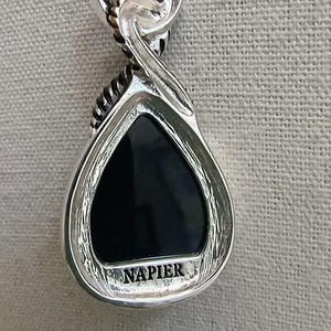 以下が含まれることがあります： 黒い涙型の石が付いたシルバーカラーのペンダント。ペンダントは、下部に「NAPIER」という言葉が刻まれた複雑なデザインです。ベイルはツイストデザインです。