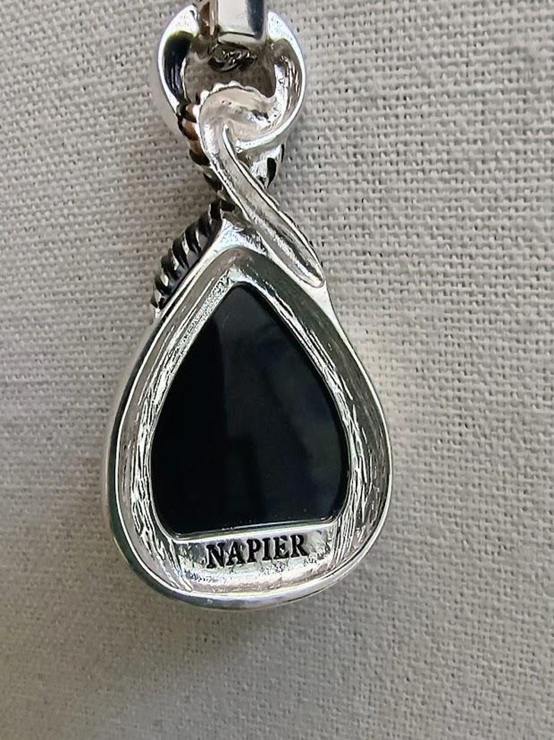 以下が含まれることがあります： 黒い涙型の石が付いたシルバーカラーのペンダント。ペンダントは、下部に「NAPIER」という言葉が刻まれた複雑なデザインです。ベイルはツイストデザインです。