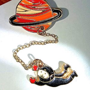 May include: A whimsical enamel pin featuring a planet with red, orange, and white stripes, a black ring, and a gold chain connected to an astronaut in a black suit with red gloves. The astronaut is floating in space.