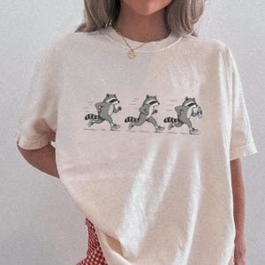 May include: A cream-colored short-sleeve t-shirt featuring a graphic print of three grey raccoons in a sequence of running motions. The raccoons are depicted with striped tails and are wearing sneakers, with the third one holding a water bottle.