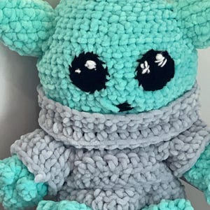 May include: A handmade crochet doll resembling a well-known character. The doll has a light teal head and limbs, large black eyes, and a grey sweater. The soft toy is designed to be cuddly, appealing to fans of the franchise.