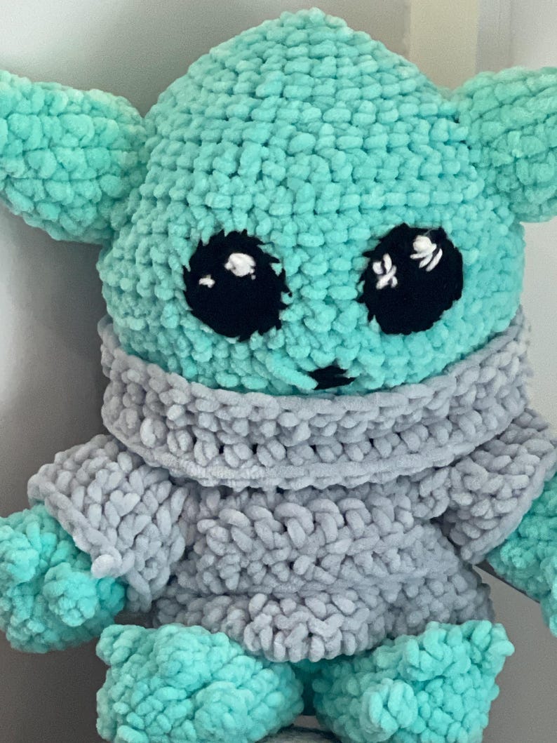 May include: A handmade crochet doll resembling a well-known character. The doll has a light teal head and limbs, large black eyes, and a grey sweater. The soft toy is designed to be cuddly, appealing to fans of the franchise.