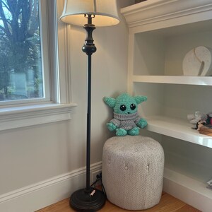 May include: A crocheted green and grey doll sits on a small, upholstered ottoman. A black floor lamp with a beige lampshade stands nearby. The room has a window and a white bookshelf.