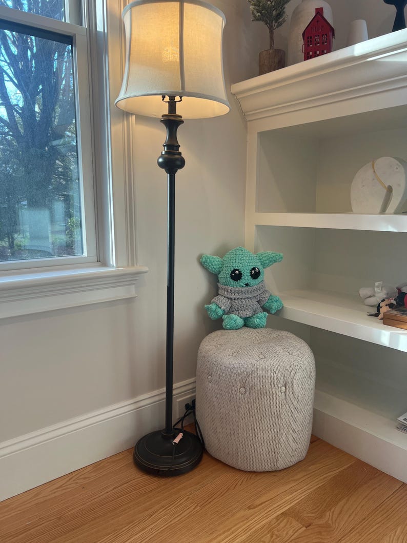 May include: A crocheted green and grey doll sits on a small, upholstered ottoman. A black floor lamp with a beige lampshade stands nearby. The room has a window and a white bookshelf.