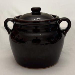 May include: A dark brown ceramic bean pot with a glossy finish, featuring two curved side handles and a matching removable lid with a central knob. The pot has a rounded, bulbous body shape and a smooth, reflective surface throughout the exterior.