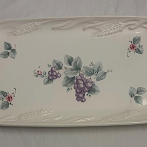 May include: A rectangular off-white ceramic serving tray featuring a central illustration of purple grapes and green leaves. The rim is decorated with a raised, embossed wheat stalk pattern, and small floral accents are placed in the corners.