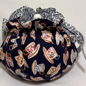 May include: A white ceramic teapot with a navy blue tea cozy. The cozy is patterned with teacups and floral designs in pink, white, and gold. The top of the cozy is a light blue floral pattern.