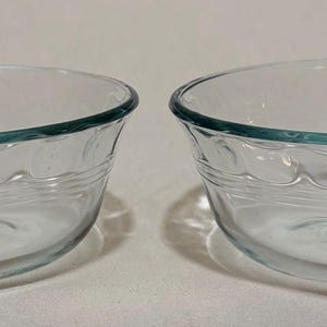 May include: Two clear glass bowls with a scalloped design around the middle and a teal rim. The bowls are oval-shaped and sit side-by-side on a white surface. The bowls are empty.
