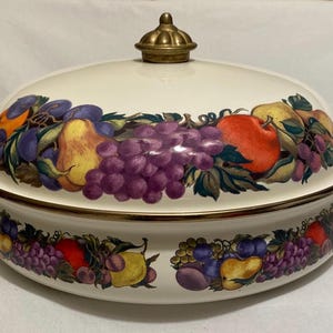 May include: Cream-colored enamel casserole dish with a lid, adorned with a vibrant fruit design featuring apples, grapes, and pears. The dish has brass-colored handles and a matching knob on the lid, perfect for serving. A decorative kitchenware item.