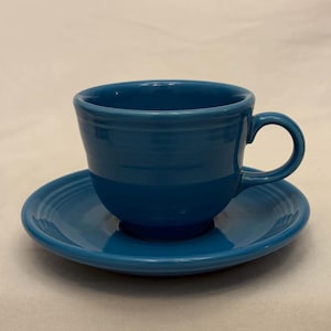 May include: A teal-colored teacup and saucer set. The cup has a rounded shape with a handle, and the saucer is circular with concentric rings. The set is made of ceramic and has a glossy finish. The background is a plain, off-white surface.