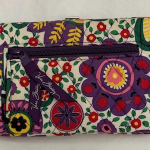 May include: A quilted wallet with a floral pattern in purple, red, yellow, and green on a cream background. It has a purple zipper and a matching pull tab with the text "Vera Bradley".