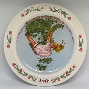 May include: A white ceramic plate with a floral border and a picture of a girl on a swing. The plate says "A certified plate in the Charles M. Schulz only Series" and "Weber Calendar Collection." The month of May is written at the bottom of the plate.