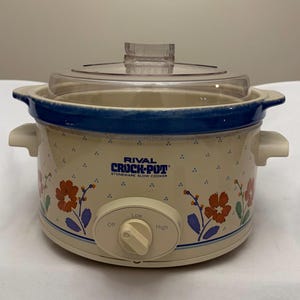 May include: A Rival Crock-Pot stoneware slow cooker with a cream-coloured base featuring a blue floral pattern and small blue dots. It has a clear glass lid, two side handles, and a front control knob with Off, Low, and High settings printed.