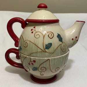 May include: A cream-colored ceramic teapot and teacup set with red accents. The teapot has a lid and a handle, and the teacup is integrated into the base. The set is decorated with green and brown floral patterns and red berries.