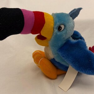 Op de afbeelding: Een pluche toekan vogel speelgoed met een grote zwarte, roze, rode en gele snavel. De vogel heeft blauwe en witte veren, oranje poten en een klein label. Het speelgoed is ontworpen om rechtop te zitten.