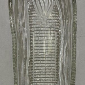 May include: Clear glass corn-shaped dish. The dish has a detailed design mimicking the texture of corn on the cob. The edges are gently curved, and the overall shape is elongated, perfect for serving corn.