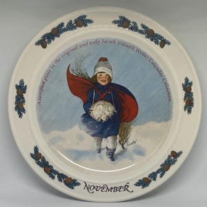 May include: A white ceramic plate with a blue and green border featuring pine cones and branches. The center of the plate has a colorful illustration of a girl in a red coat and blue dress holding a snowball. The text "A certified plate in the original and only Sarah Silwell Weber Calendar Collection." is written around the illustration. The text "NOVEMBER" is written at the bottom of the plate.