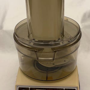 Peut inclure: Un robot culinaire General Electric vintage beige et marron. L'appareil comprend un bol en plastique transparent et un grand récipient beige sur le dessus. La base comporte des boutons de commande étiquetés "Off", "On" et "Pulse On". Le logo GE et "Food Processor" sont également visibles.
